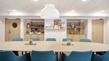 Un salón de conferencias con una gran mesa ovalada, sillas azules, una lámpara colgante blanca y una kitchenette con estantes arriba.