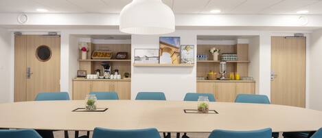 A conference room with a large oval table, blue chairs, a white pendant light, and a kitchenette with shelves above.