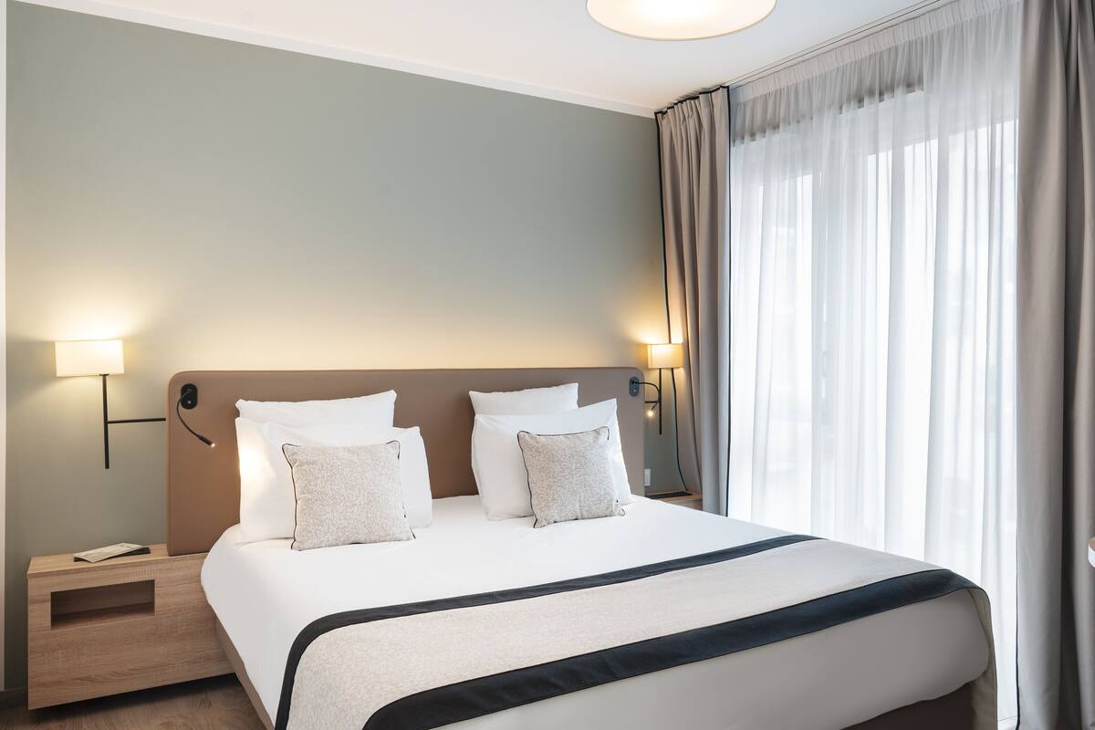 Hotelkamer met een groot bed, twee nachtkastjes, een ronde tafel en een raam met gordijnen.