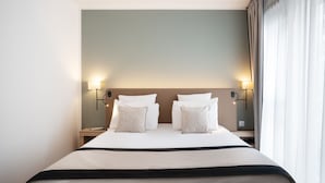 Un letto rifatto con lenzuola bianche e due cuscini decorativi, un comodino con una lampada, e vista su una finestra con tende.