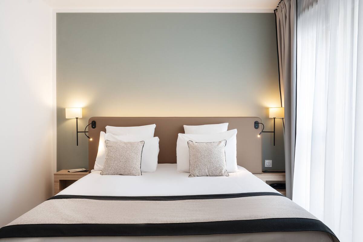 Een net opgemaakt bed met witte lakens en twee decoratieve kussens, een nachtkastje met een lamp, en uitzicht op een raam met gordijnen.