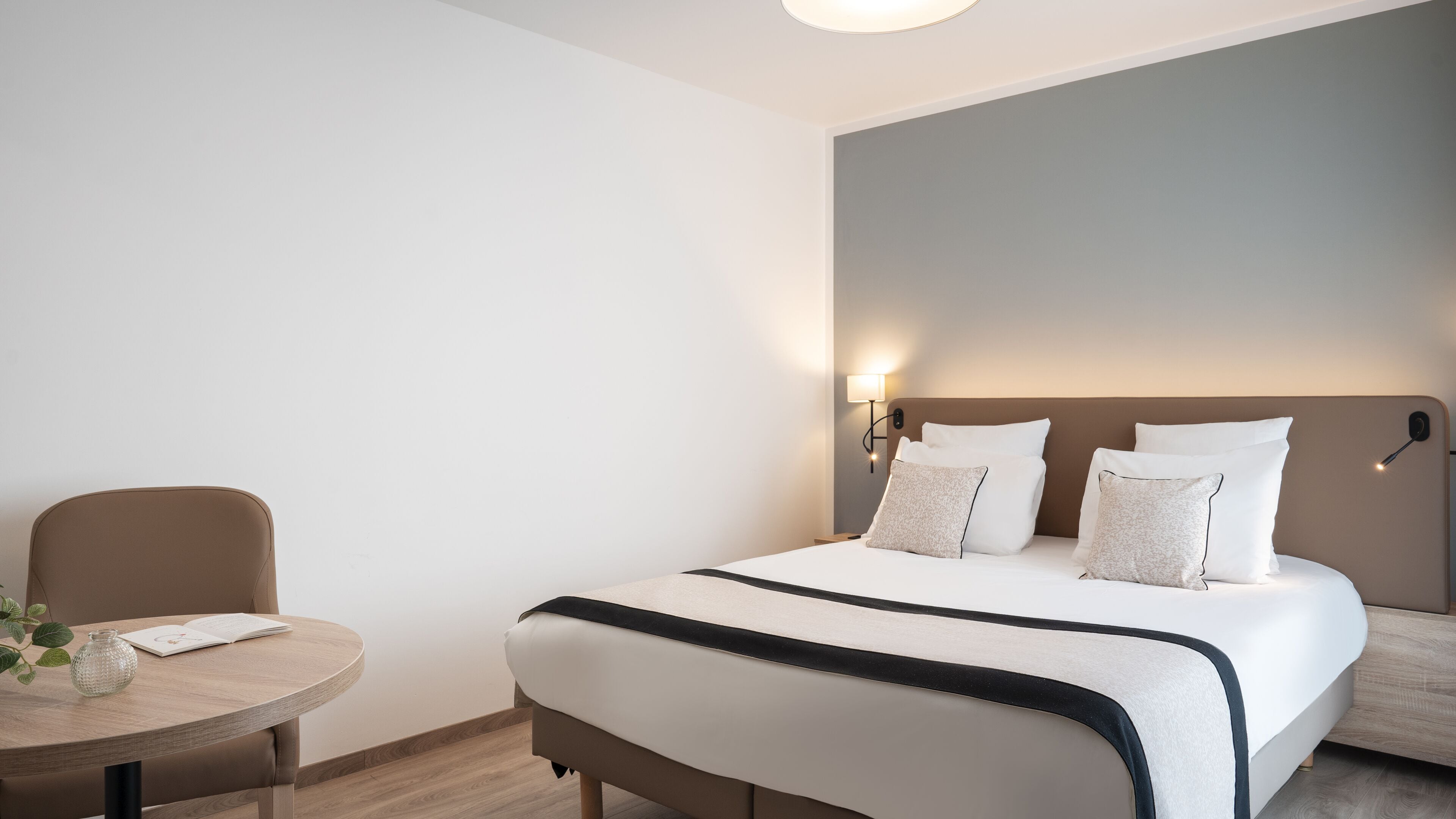 A modern hotel room with a neatly made bed, a round wooden table with a book and a vase, and a wall-mounted lamp.