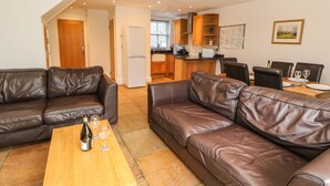 A living room with a brown leather sofa, a wooden coffee table with a bottle and glasses, and a kitchen in the background. - Brancepeth (Bishop Auckland)
