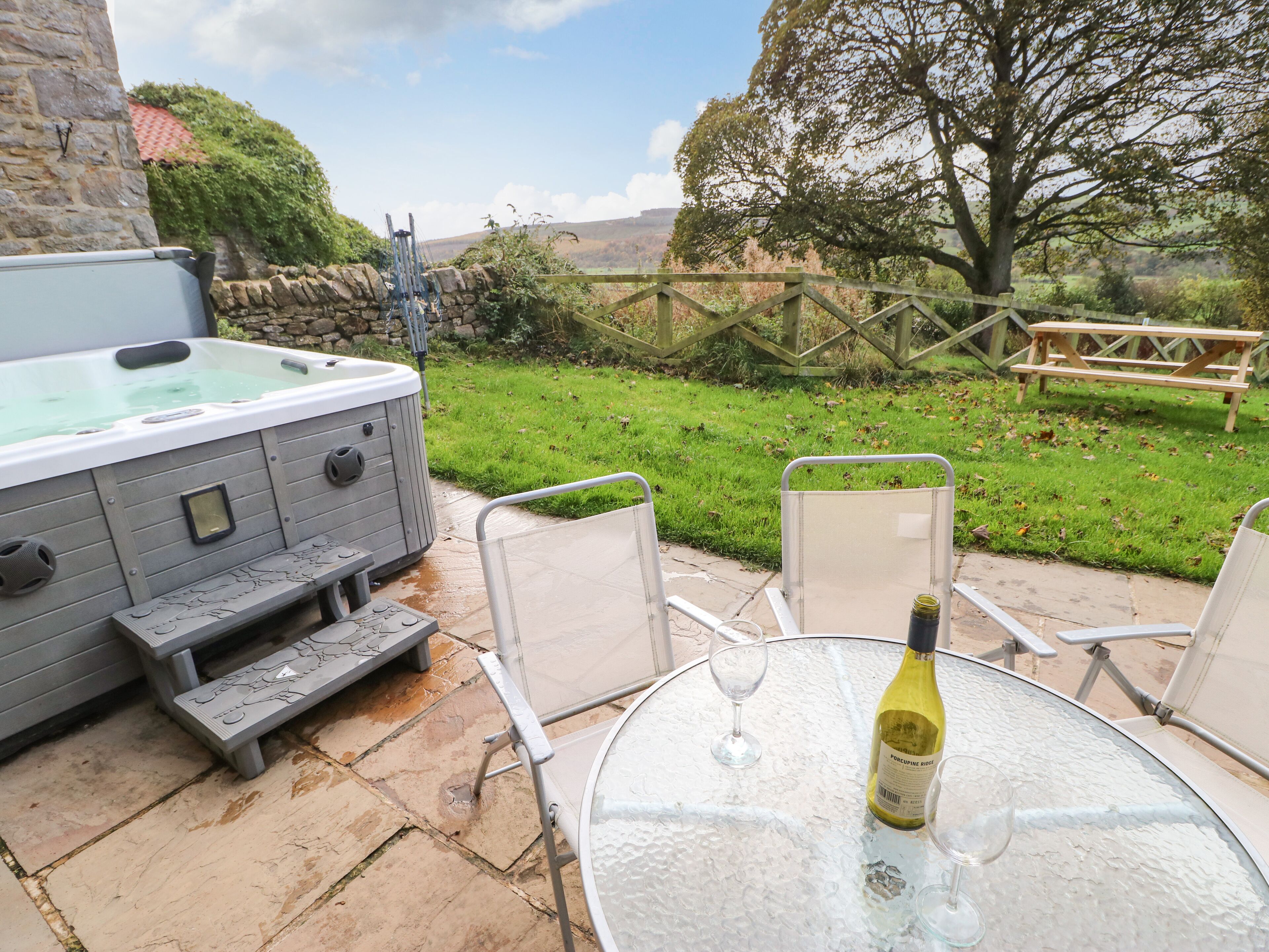 A hot tub with a stone wall backdrop, a wooden picnic table, and a glass table with a bottle and glass.