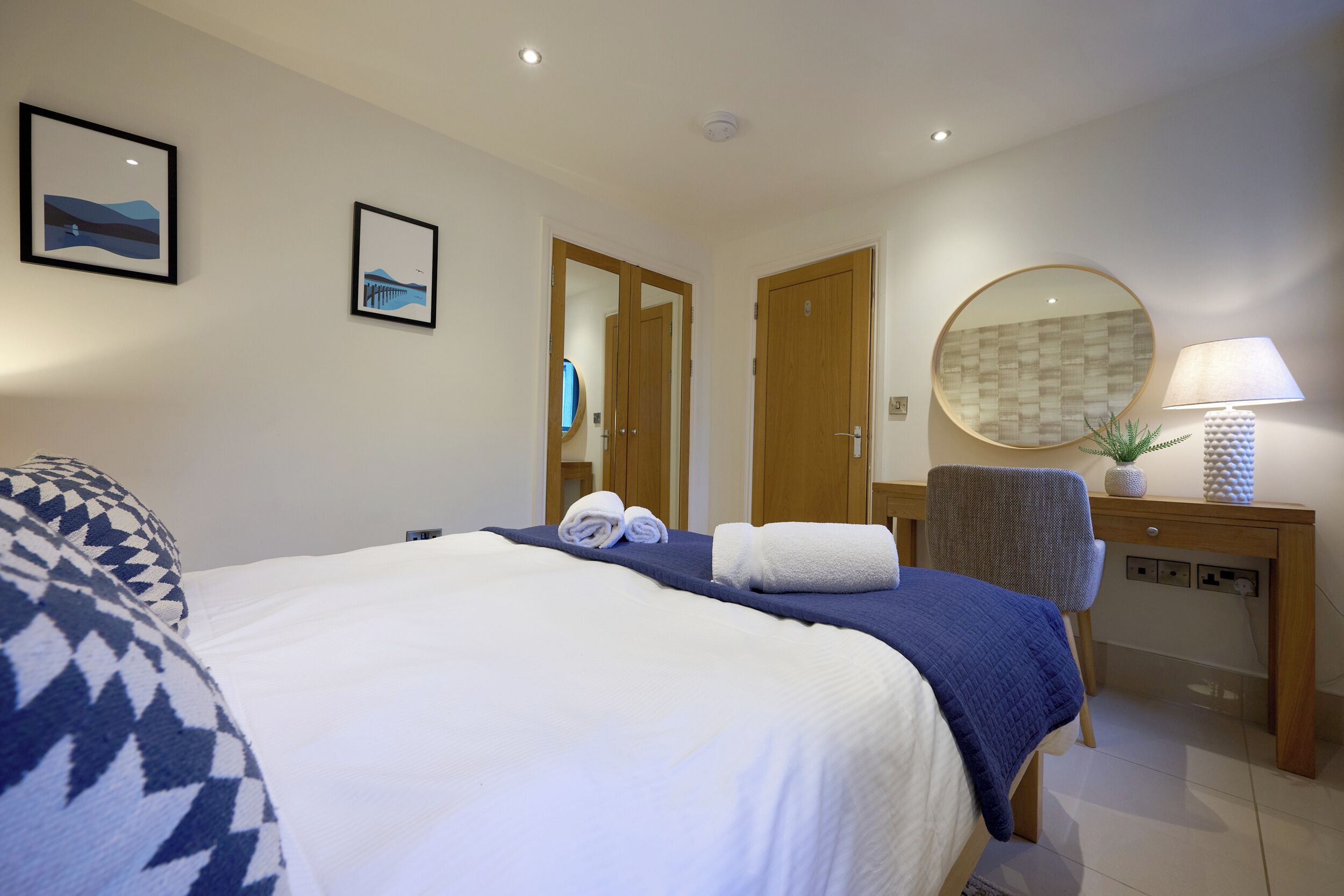 A hotel room with a bed, a desk, a chair, a round mirror, and two framed pictures on the wall.