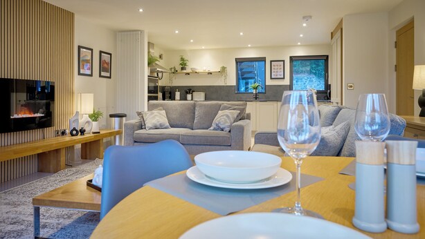 Un salon moderne avec une cheminée, des canapés gris, une table à manger avec de l’assiette et des verres, et une cuisine en arrière-plan.