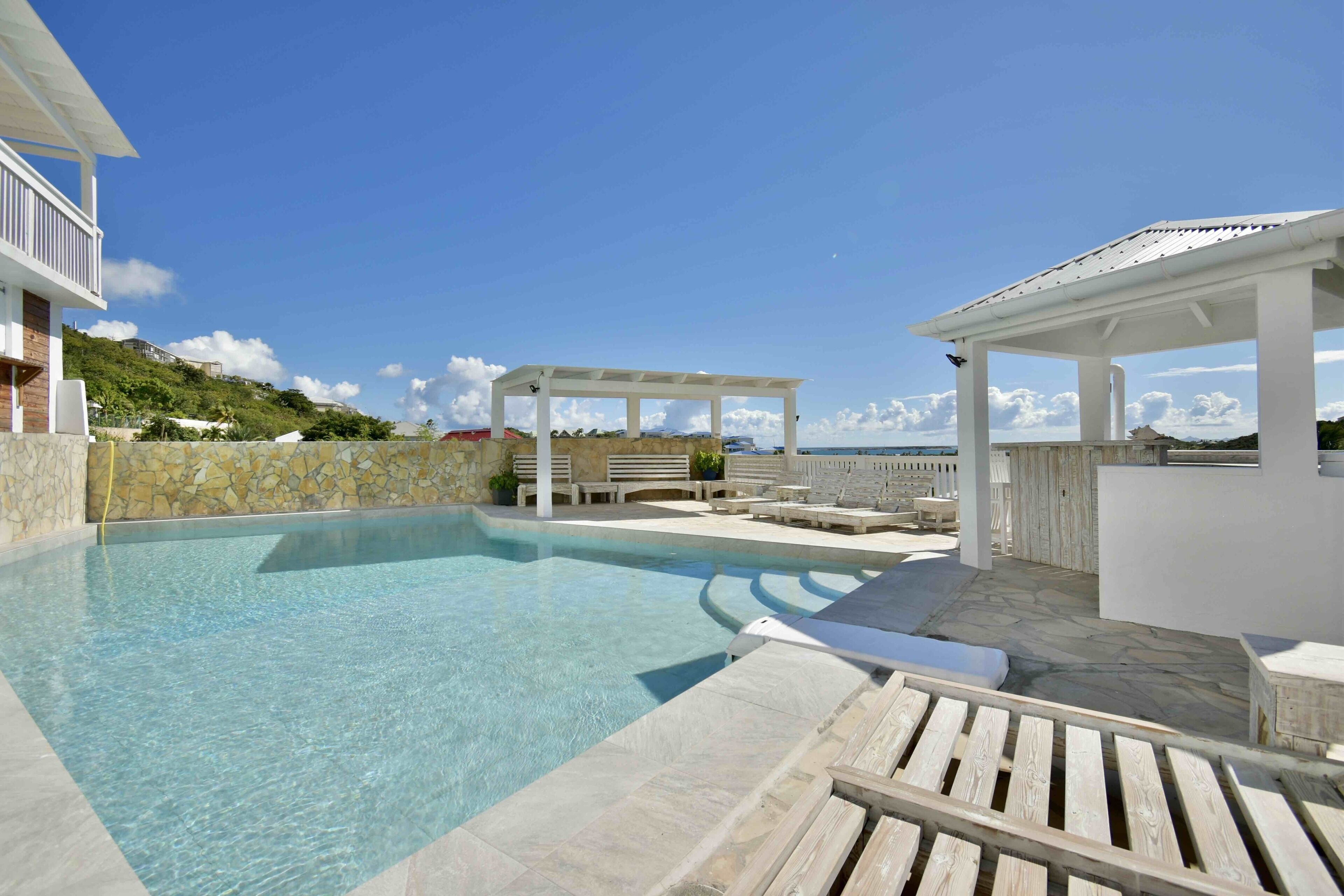 A clear swimming pool with lounge chairs and a pergola, surrounded by stone walls and a view of the sea.