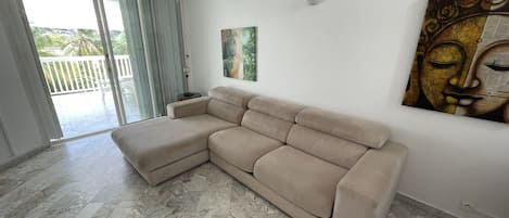 A beige sectional sofa in a room with a marble floor, a painting on the wall, and a view through a sliding glass door to an outdoor area with a white fence and palm trees.