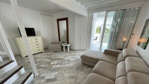 Een ruime woonkamer met een beige bank, een klein rond tafeltje en een televisie op een witte kast.