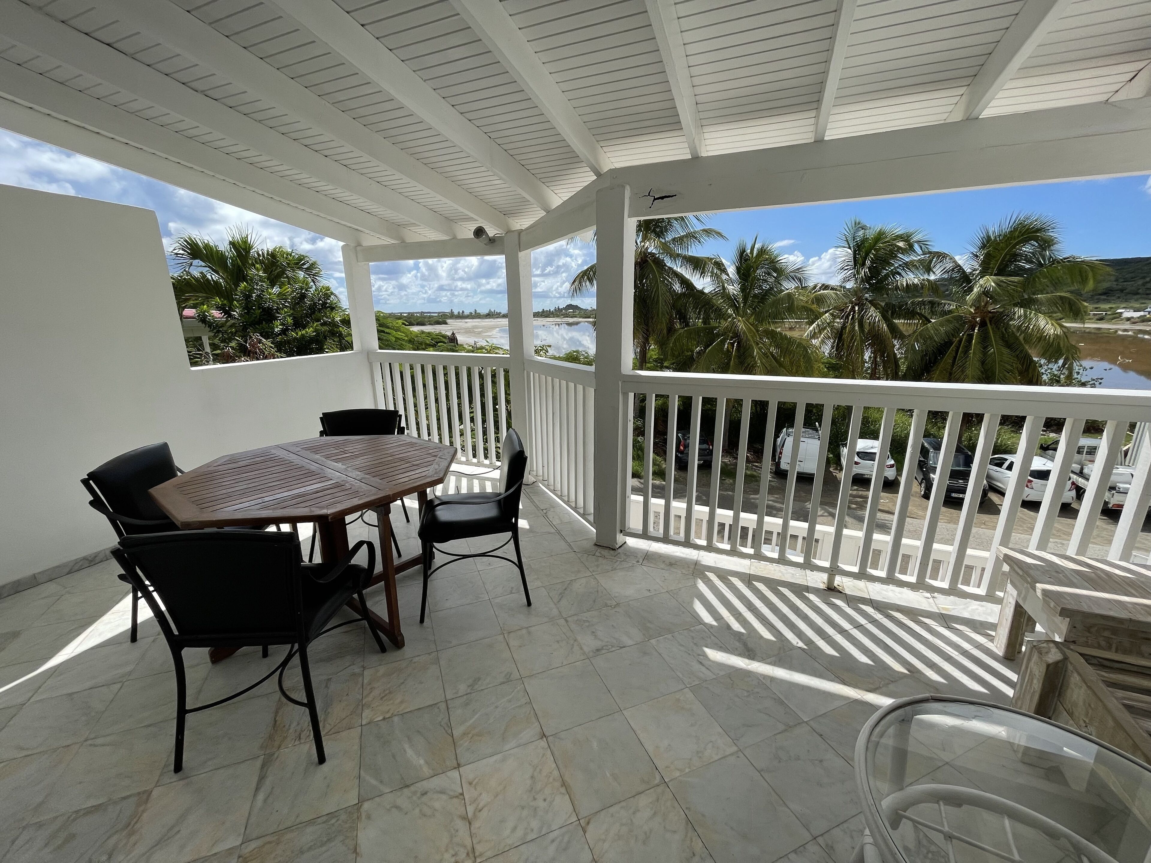 A covered outdoor area with a table and chairs, a white railing, and a view of palm trees and a beach.