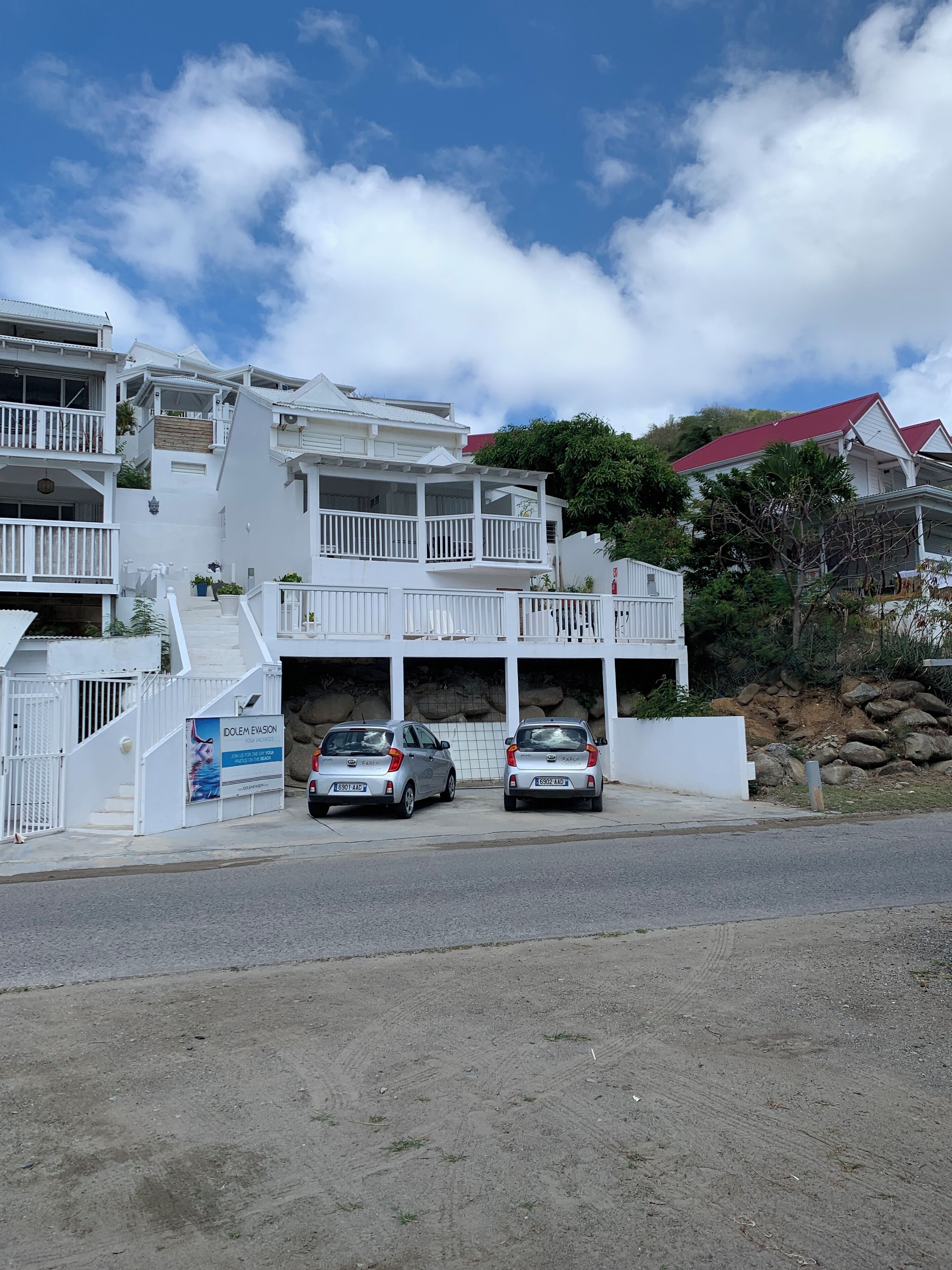 Un bâtiment blanc doté de nombreux balcons et d’un toit rouge, deux voitures garées et une enseigne publicitaire pour un complexe hôtelier en bord de mer.