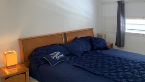 Een slaapkamer met een houten hoofdbord, een bed met een donkerblauwe spreidekker en kussens, een bedlamp en een raam met gordijnen.