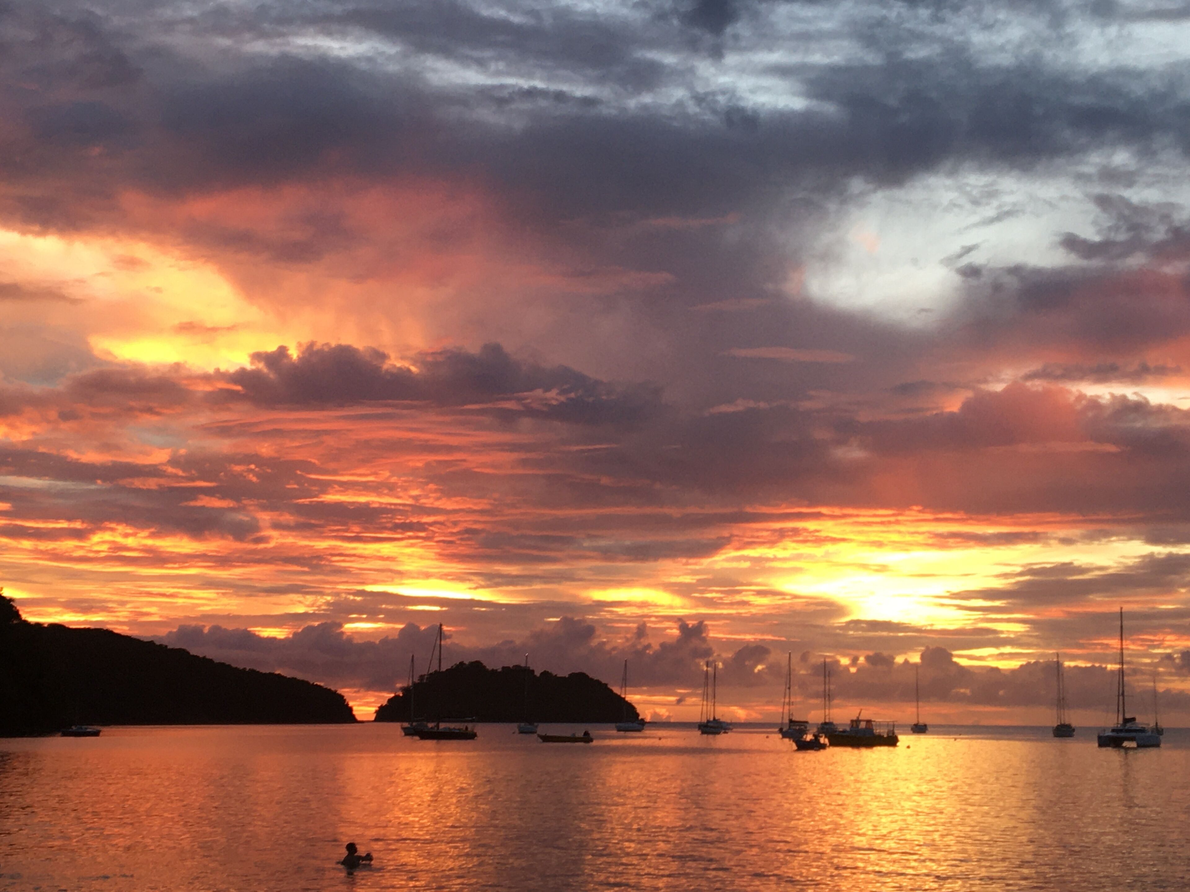 A sunset over a calm sea with several boats anchored, a small island in the distance, and a person swimming in the water.