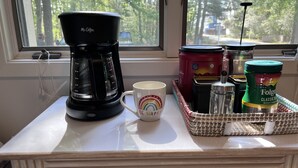 A kitchen counter with a coffee maker, a mug, a basket of tea bags, and a jar of Folgers coffee.