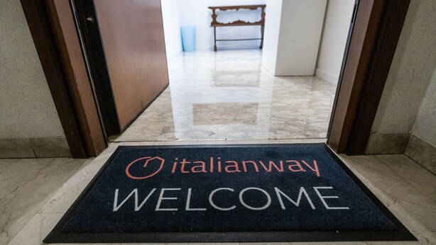Un tappetino di benvenuto con la scritta "Italianway" e "BENVENUTO", che conduce a una stanza con una panca e un cestino.