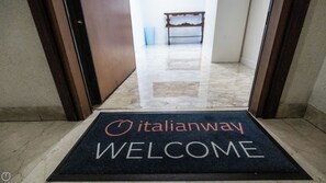 A welcome mat with 'Italianway' and 'WELCOME' text, leading to a hallway with a bench and a trash bin.