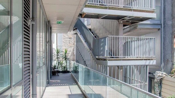 Ein moderner Gebäudekorridor mit Glastrennvorrichtungen, einem Backsteinboden und Blick auf ein mehrgeschossiges Gebäude mit Balkonen.