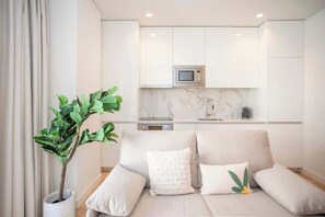 A modern living room with a sofa, a potted plant, and a kitchen area with white cabinets and a microwave. - Liiiving in Porto - City Design Apartment G (Porto)