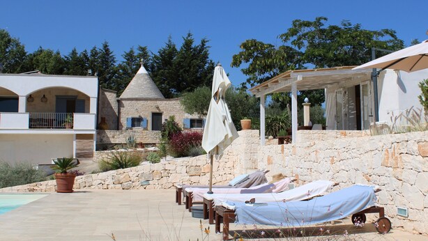 A stone-walled pool area with lounge chairs and umbrellas, a small building with a conical roof, and a white building with a balcony. - Stylishly restored trullo with private pool and stunning views (Martina Franca)