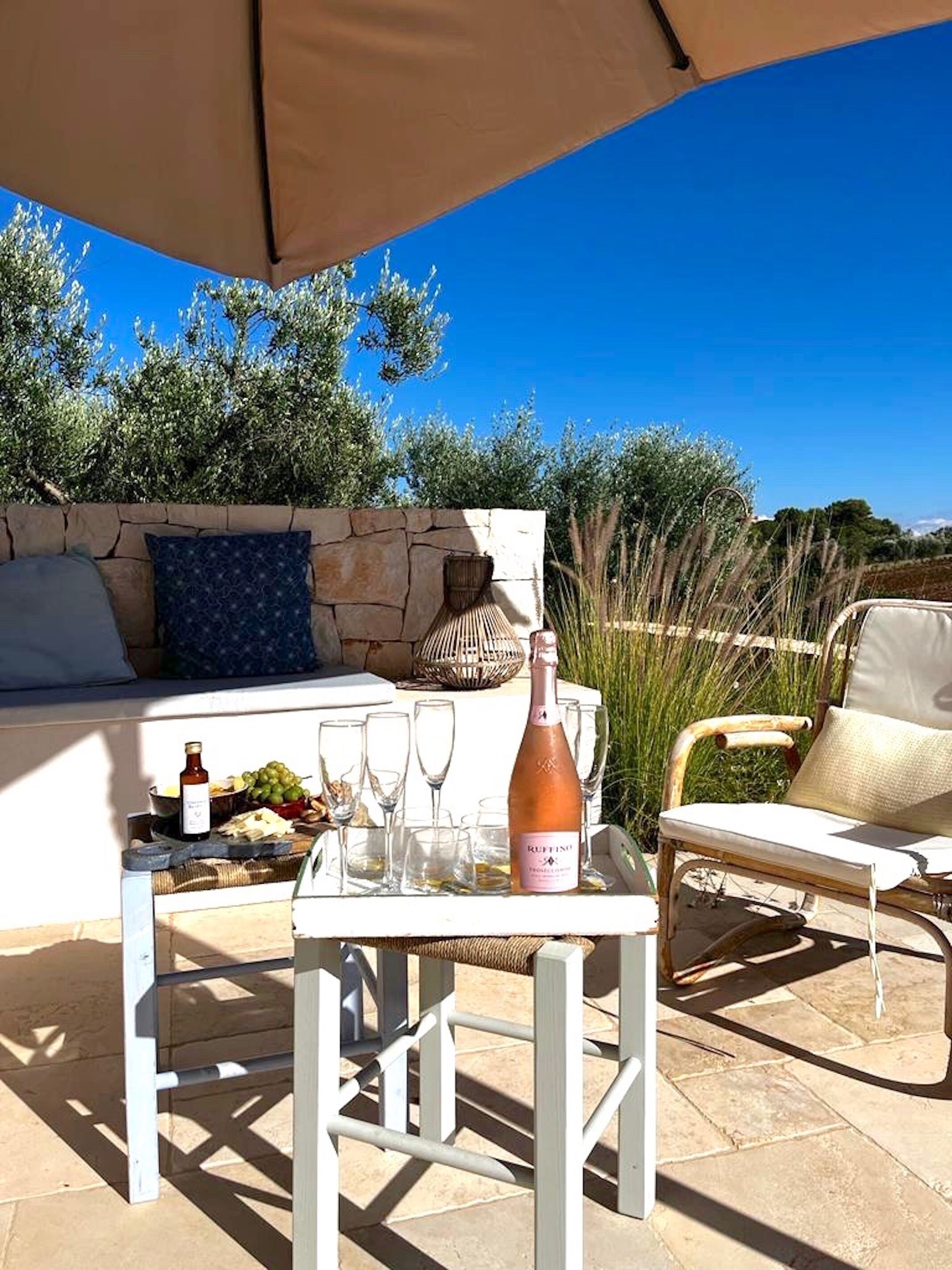 An outdoor seating area with a table, chairs, a bottle of wine, and a stone wall with greenery in the background.