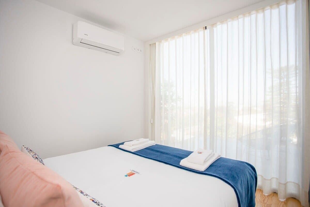 A hotel room with a bed, a window with sheer curtains, and an air conditioning unit on the wall.