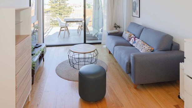 Ein modernes Wohnzimmer mit Holzboden, einem grauen Sofa, einem kleinen runden Tisch und einer Schiebeglastür, die zu einem Außenbereich führt.