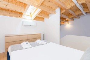 Een moderne slaapkamer met een schuine kap, houten balken, een bed met witte lakens, een nachtkastje en een aan de muur bevestigde airconditioner.