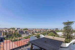 Een balkon met een houten tafel en witte stoelen, met uitzicht op een stadsgezicht met gebouwen en een heldere lucht.