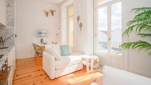 A bright living room with a white sofa, a small table, and a dining area with a white table and chairs. There is a large window providing natural light and a view of the outside. - Liiiving in Porto - Downtown Balcony Studio (Porto)