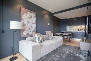 A modern living room with a grey sofa, a large abstract painting, a floor lamp, and a kitchen area in the background.
