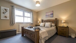 A bedroom with a wooden bed, a nightstand, a bedside table, a radiator, a window with curtains, and a painting on the wall. - Harry s - 2 Bedroom Holiday Home - Tenby (Tenby)