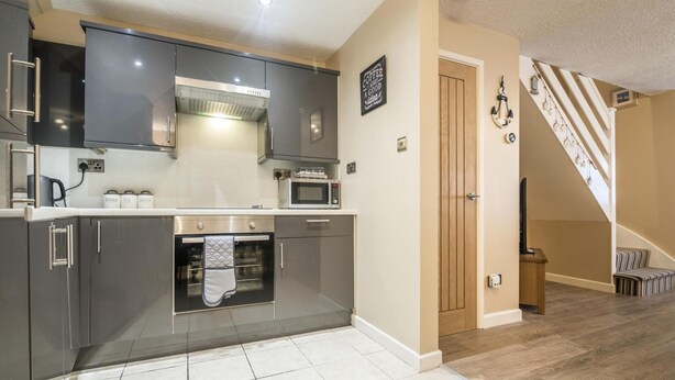 A modern kitchen with dark cabinets, a stainless steel range hood, a microwave, and a built-in oven. There's a staircase leading to a lofted area with a view. - Harry s - 2 Bedroom Holiday Home - Tenby (Tenby)