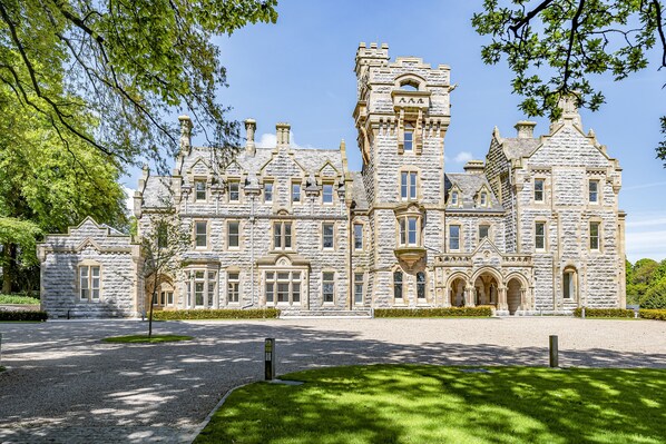 A large stone building with multiple towers and arched windows, surrounded by a well-maintained lawn and gravel paths.