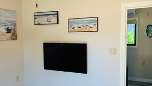 Ein Zimmer mit einem an der Wand befestigten Flachbildfernseher, drei gerahmten Kunstwerken mit Strandansichten und einer Tür zu einem weiteren Raum.