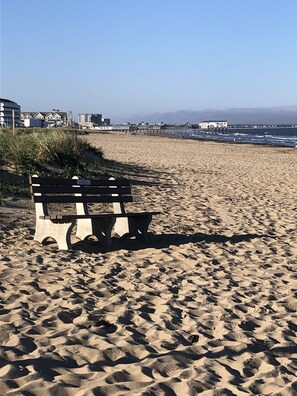 A beach with a wooden bench, sandy dunes, and buildings in the background. - Ocean view, fully renovated beach condo!!! Steps to the sand, sleeps 6 (Old Orchard Beach)