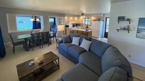 A modern living room with a gray sofa, a wooden coffee table, and a dining area with a table and chairs. A kitchen with wooden cabinets and a refrigerator is visible in the background. - “Stay Anchored” Lake Tippecanoe channel front with pier and great lake views (Leesburg)