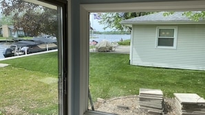 A view from inside a room with a large window overlooking a grassy yard, a dock with a covered boat, and a small house across the water. - “Stay Anchored” Lake Tippecanoe channel front with pier and great lake views (Leesburg)