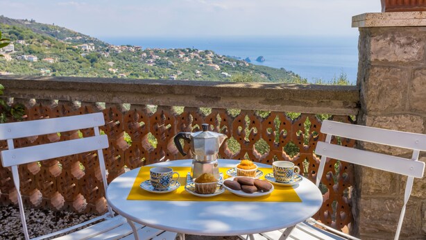 Un tavolo apparecchiato con una caffettiera, cupcakes e piattini di biscotti su un balcone con vista su una cittadina costiera.