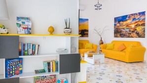 Un soggiorno con un divano giallo, una libreria piena di giochi da tavolo e un quadro appeso al muro.