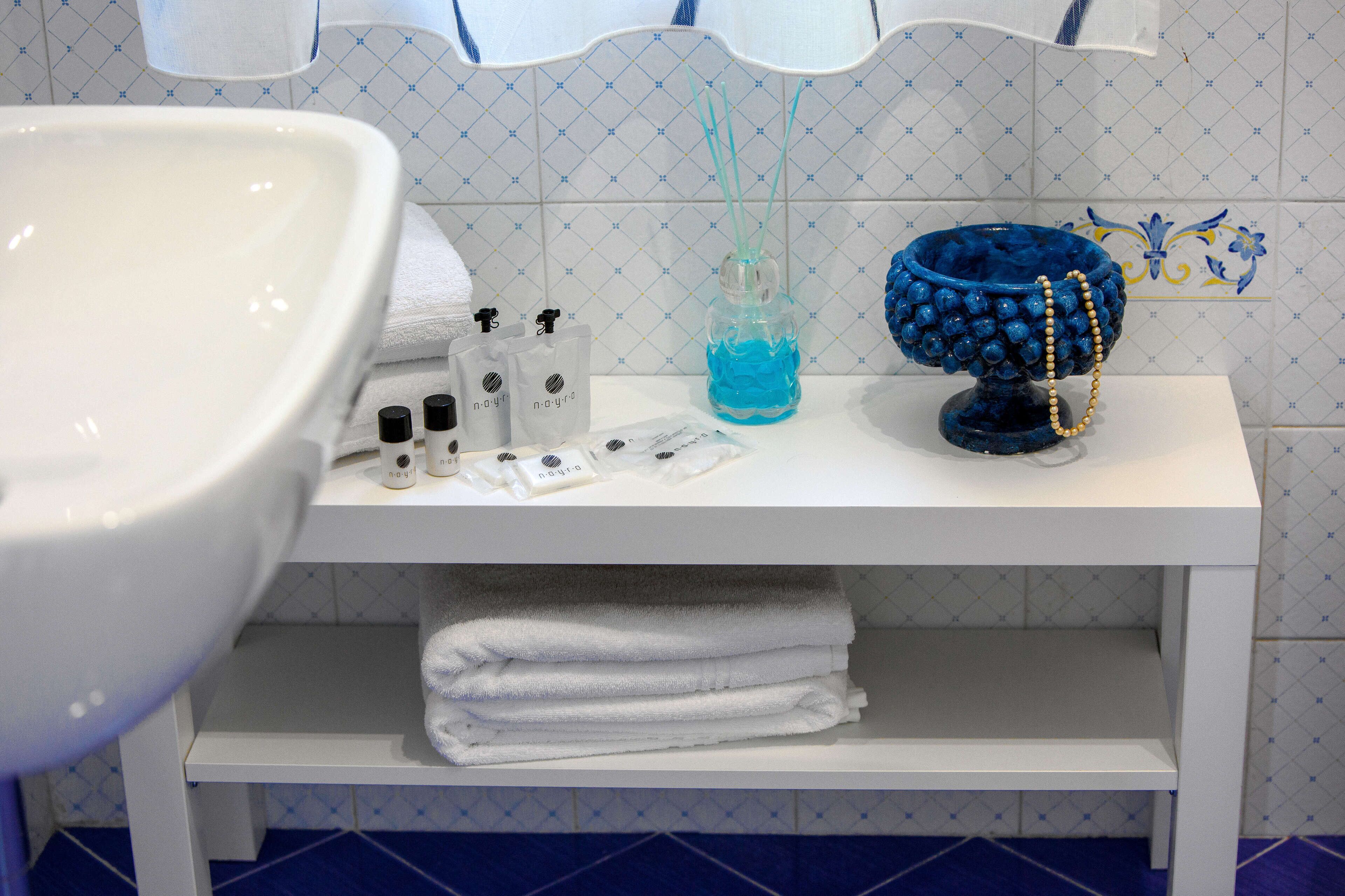 Un baño con un lavamanos blanco, un estante blanco con toallas dobladas, un cuenco decorativo azul y una botella azul con un cordón amarillo.