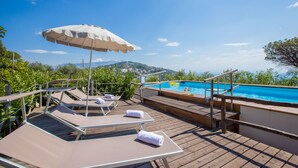 Un terrazzo in legno con poltrona relax e ombrellone, affacciato su una piscina con vista sulle colline.