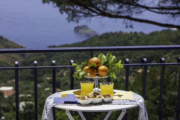 Une table avec un panier de fruits, deux verres de jus d’orange et une petite assiette de noix, sur un balcon offrant une vue panoramique sur un lac et la verdure environnante.