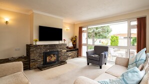 A living room with a stone fireplace, a flat-screen TV, and a large window with a view of the outdoors. - Longhouse Bungalow (Wadebridge)