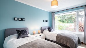 A bedroom with two beds, a window with curtains, and a hanging light fixture. - Longhouse Bungalow (Wadebridge)