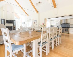 Un comedor amplio con una mesa de madera y sillas blancas, una cocina con electrodomésticos modernos, y un techo alto con vigas a la vista.