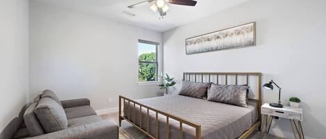 Une chambre à coucher comprenant un lit, un canapé, une petite table, un ventilateur de plafond, une fenêtre et une œuvre d’art accrochée au mur.