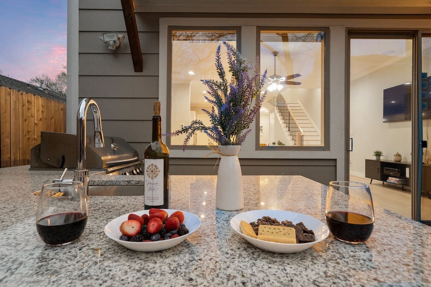 An outdoor dining area with a granite countertop, two wine glasses, a bottle of wine, and a bowl of fruit.
