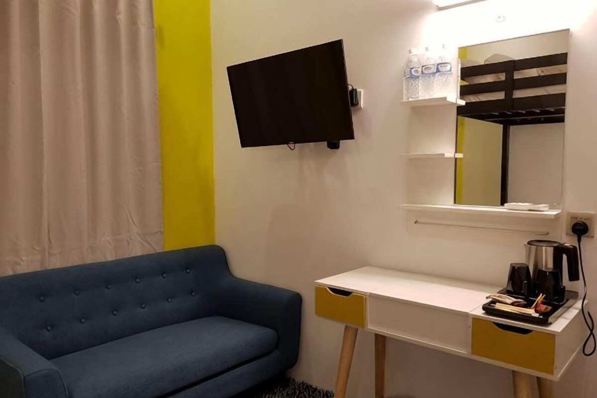 Una habitación con un sofá azul, un escritorio blanco con espejo, un televisor de pantalla plana y una cafetera.