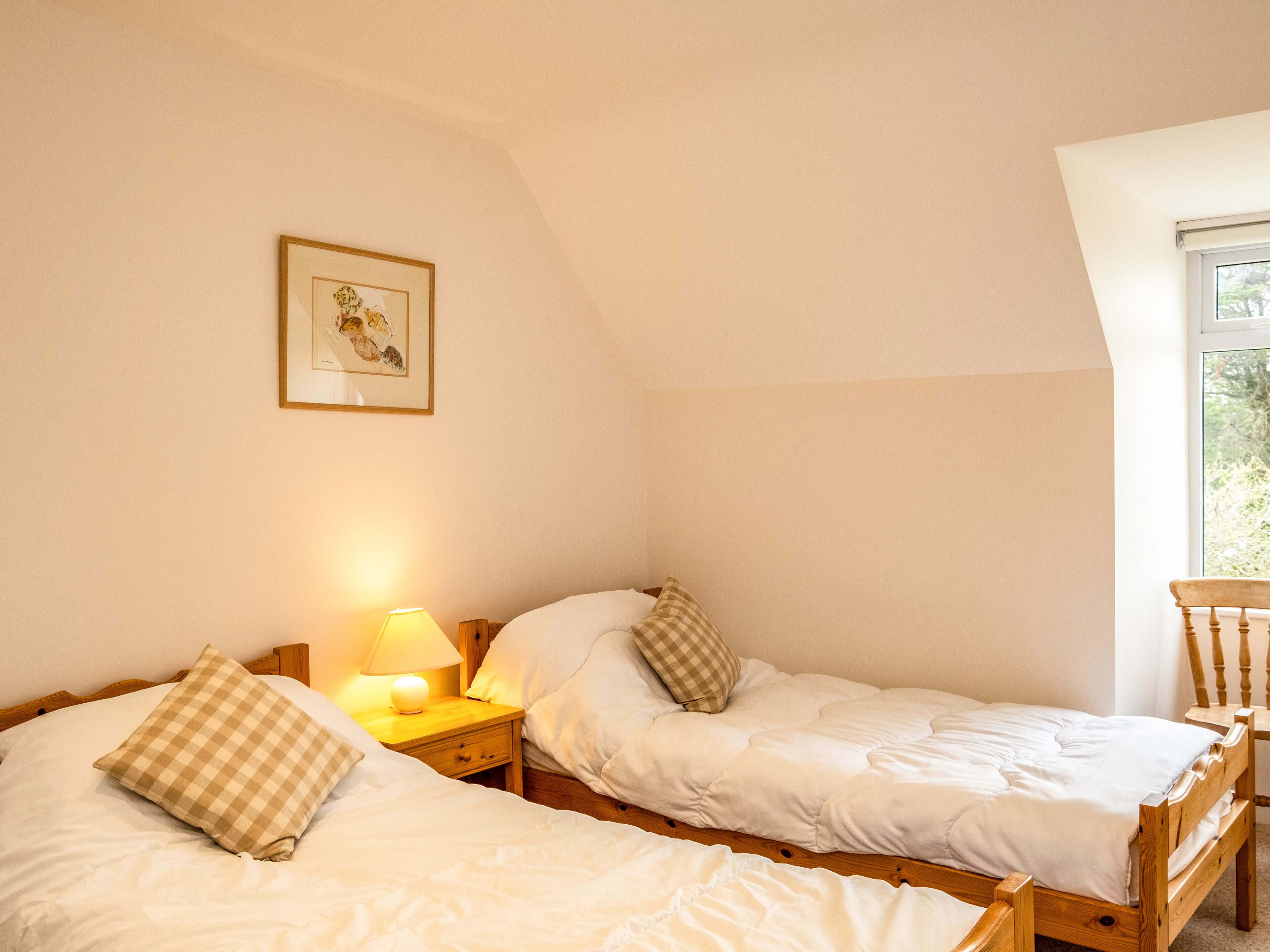 Un dormitorio con dos camas individuales, una mesita de noche con lámpara, una silla de madera y una ventana con vistas a árboles.