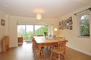 A dining area with a wooden table and chairs, a radio, and a lamp. There are framed pictures on the wall and a window with a view of greenery. - Tregillan (Wadebridge)